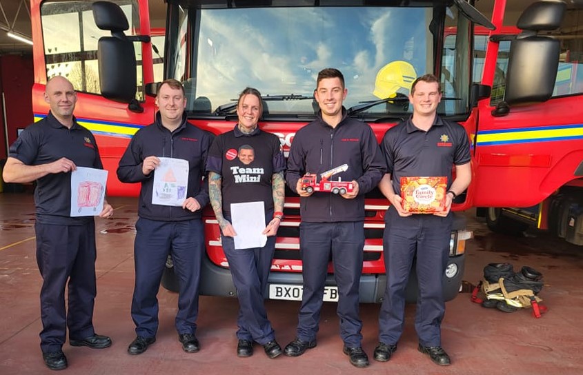 Group of smiling firefighters stand in front of an engine holding a toy fire truck, biscuits and bits of paper