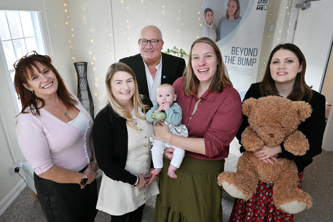 6 people (one is a little baby) in a room decorated with lights and a standing advertisement for 'Beyond the bump'. The person on the far right is holding a large brown cuddly teddy bear.