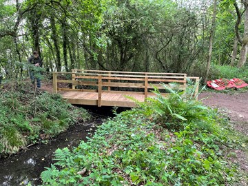 A new wooden bridge installed by volunteers at Kingsbury Water Park.  The bridge crosses a small stream and is surrounded by trees.