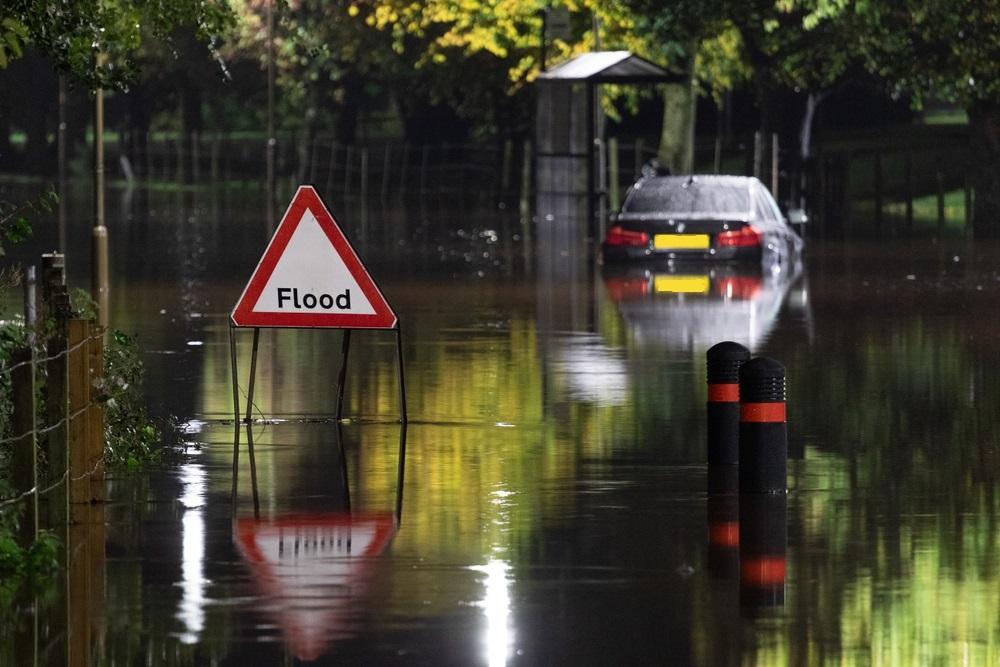 Car stuck in floodwater in background, with red and white sign reading 'Flood' in black text in foreground.
