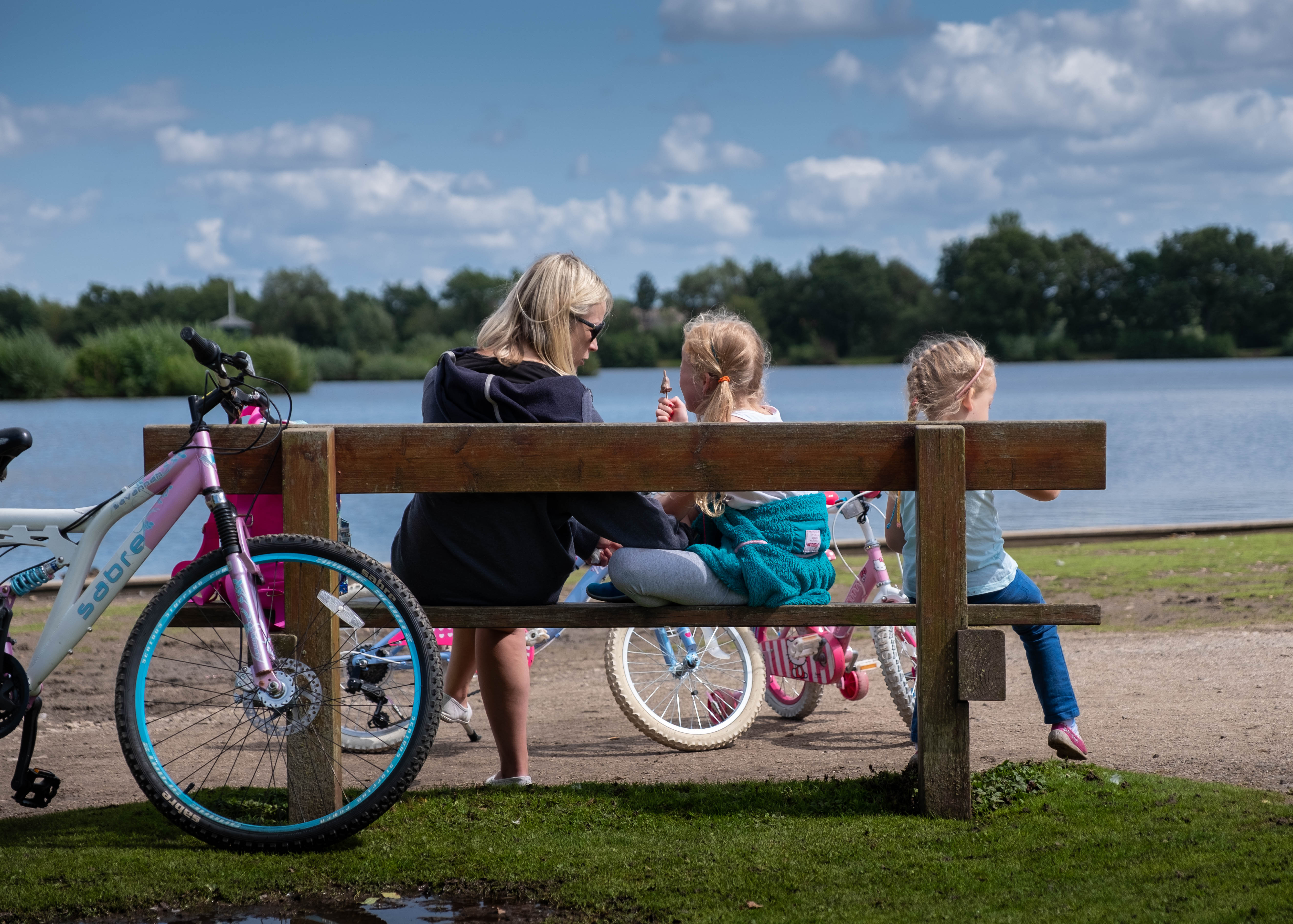 A woman sits on a bench with her two daughters. There are three bicycles in view. The bench faces a lake at Kingsbury Water Park on a clear day.