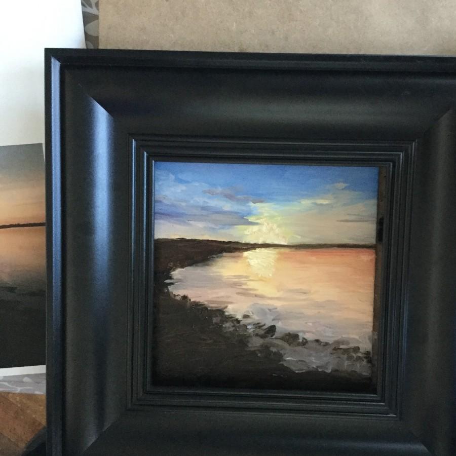A black framed painting of curved rocky lakeside with an orange and yellow sunset reflected across a lake.