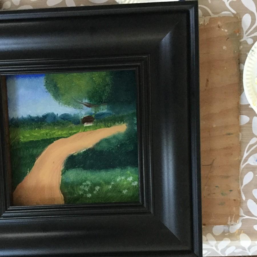 A black framed painting of a brown path surrounded by green grass under a clear blue sky. A big green tree on the right of the brown path.