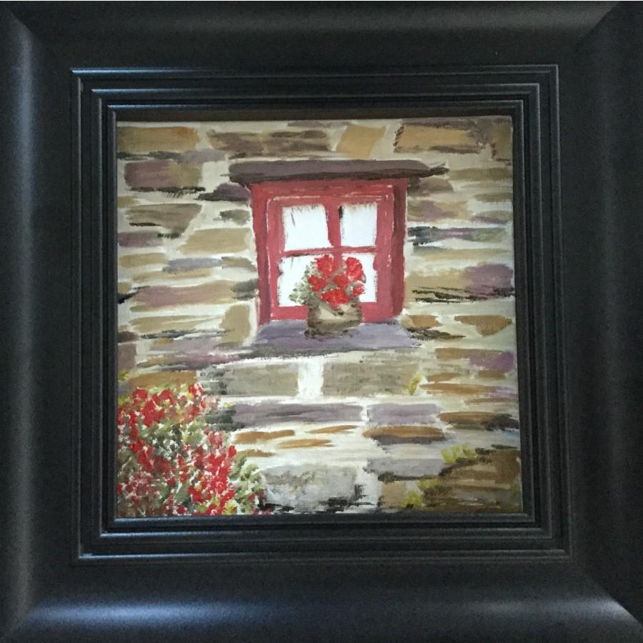 A black framed painting of a brown and grey stone house with a red window. On the window sill there is a potted plant with red flowers. Below the window sill on the left are some red flowers.