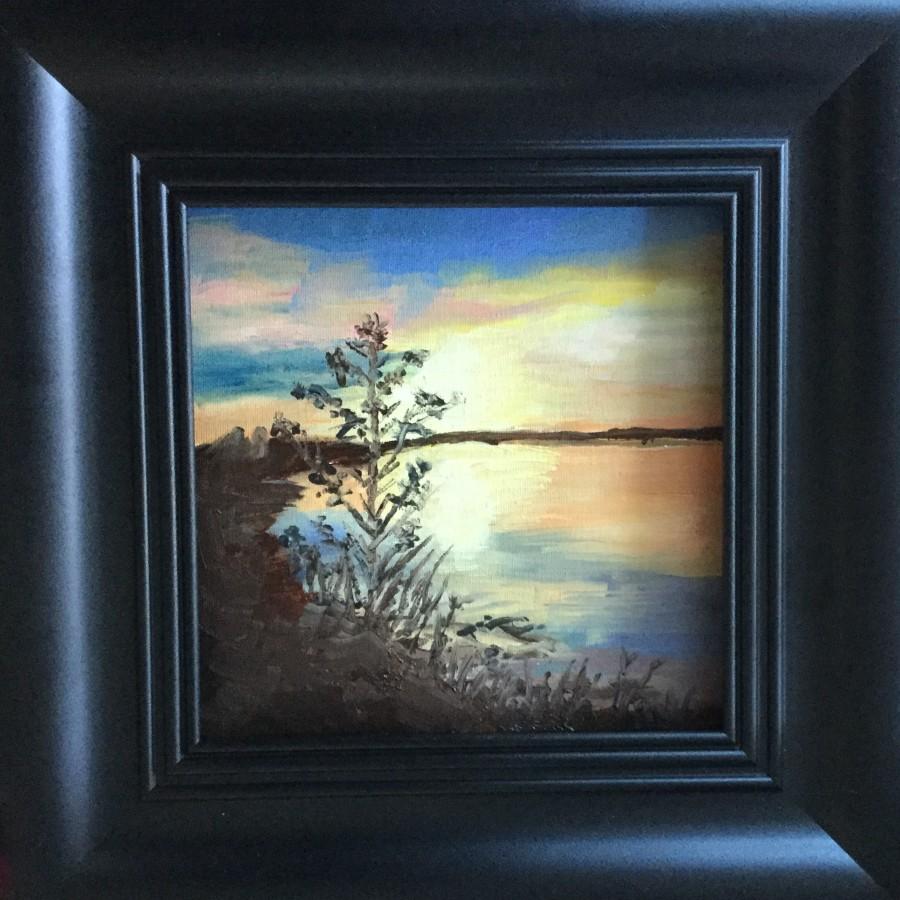 A black framed painting of an orange and yellow sunset reflected across a lake. On the left of the painting are some small tree branches.