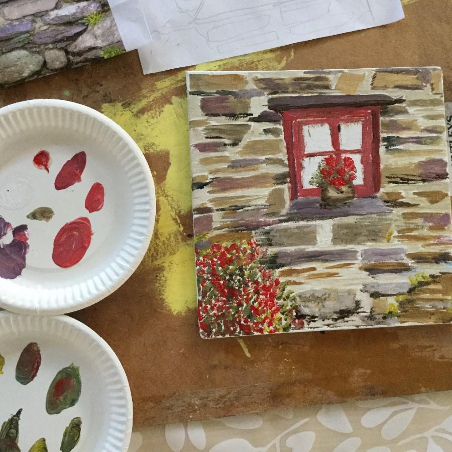 painting of a brown and grey stone house with a red window. On the window sill there is a potted plant with red flowers. Below the window sill on the left are some red flowers.