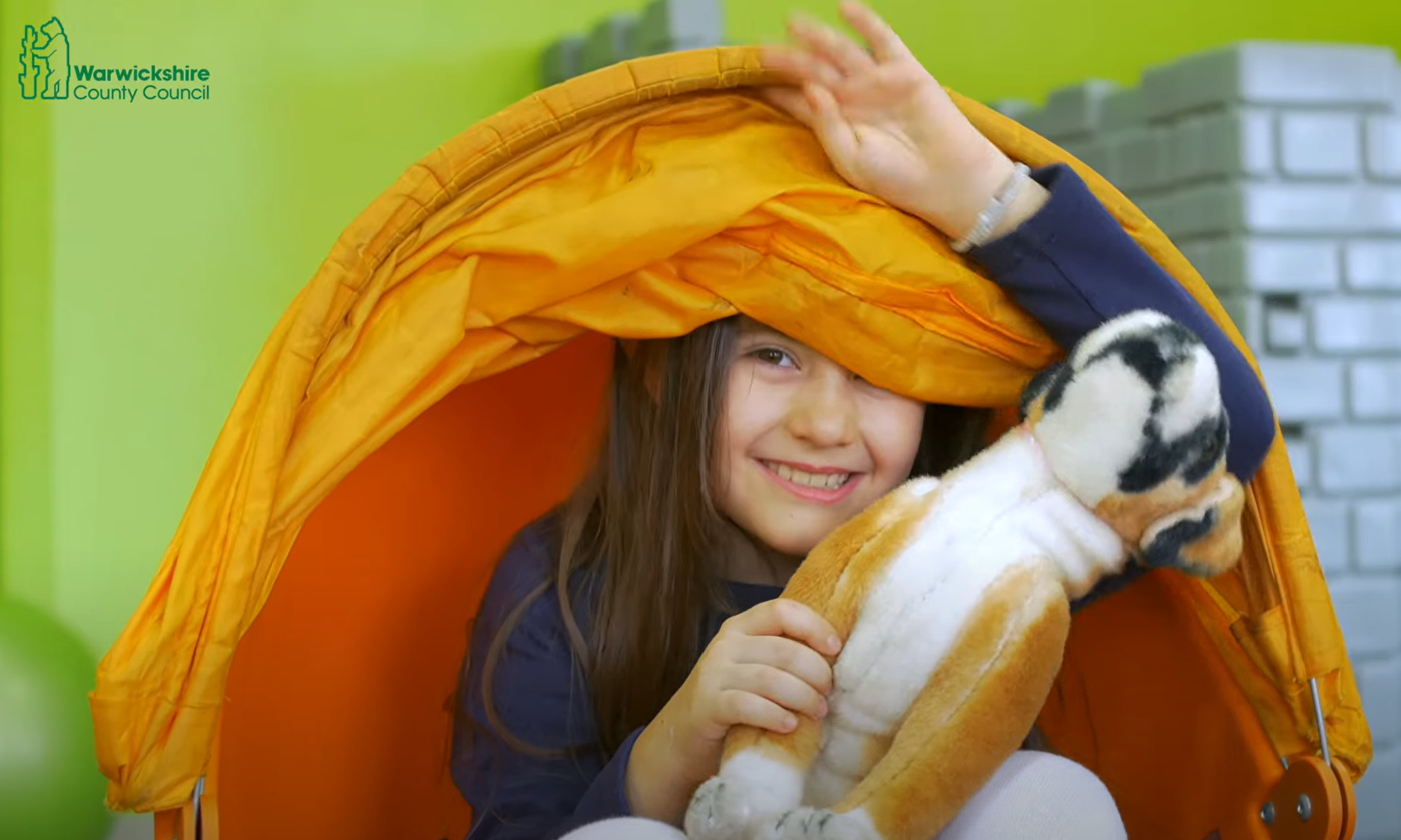 Girl smiling and sitting inside a sensory tent holding a cuddly toy and peeking out