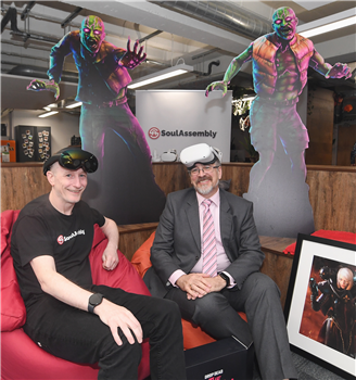 Two men wearing virtual reality headsets, sitting on a red sofa.