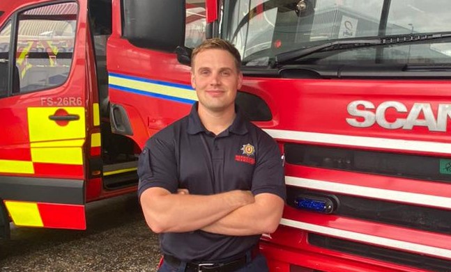 Firefighter Stephen Balestrini standing smiling with arms folded in front of two fire engines