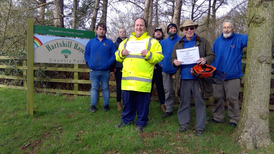 Tony Beck, Lee Charnell, (both holding certificates) and other Country Park volunteers stand by a Hartshill Hayes Country Park sign.