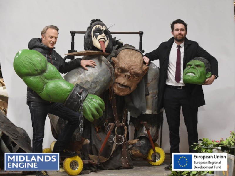 two white men holidng props and large costume items, on a hwite background. Item include a goblin mask and wearable green Hulk arms.