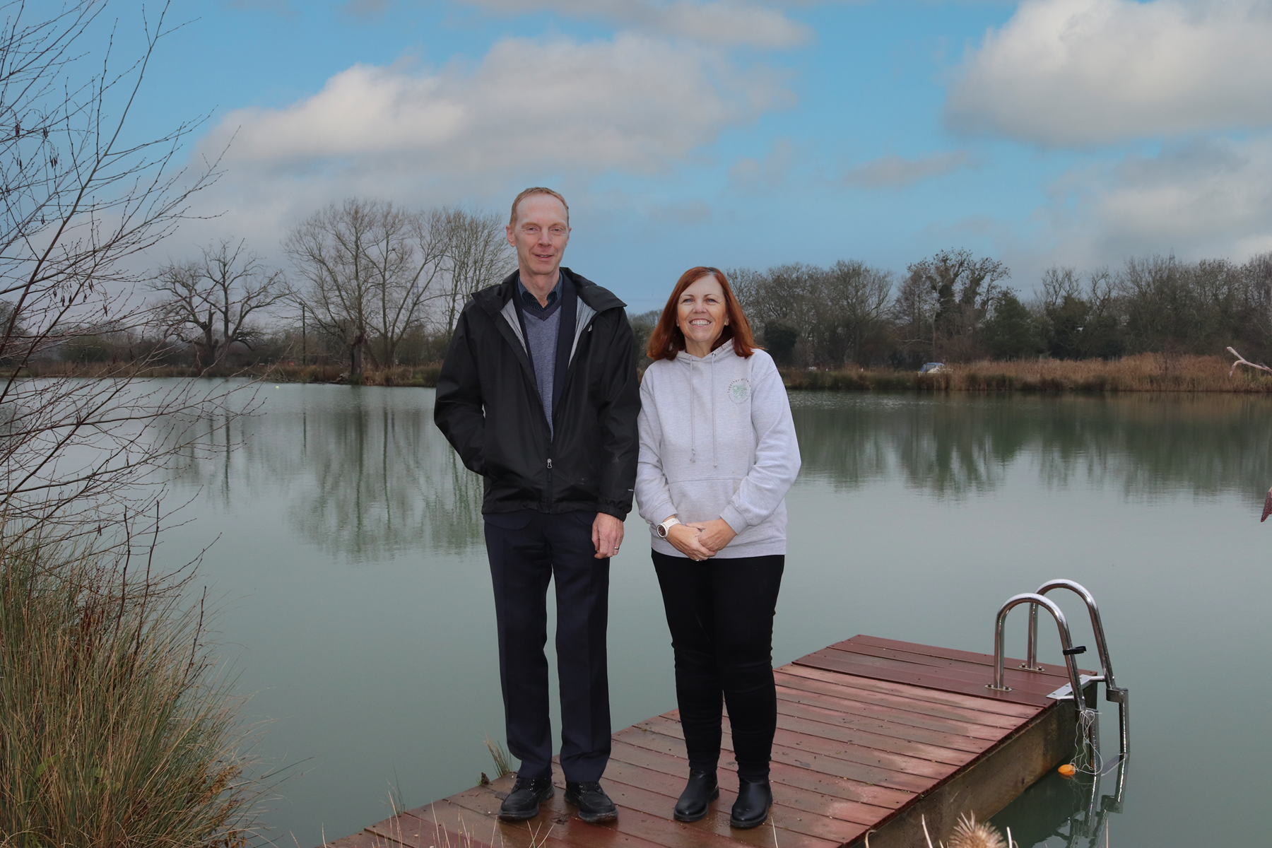 A man (left) and a woman (right) stood on a pier in the foreground, with a lake in the background.