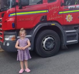Smiling child standing in front of a fire engine in a purple dress