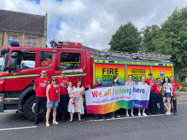 Firefighters, council workers and members of the public with a Pride wrapped fire engine and a banner that says 'We all belong here'