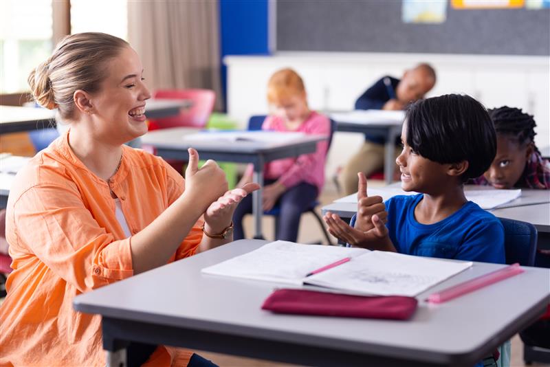 Teacher bends down next to a pupil sat at a desk in the classroom and is using hands to communicate